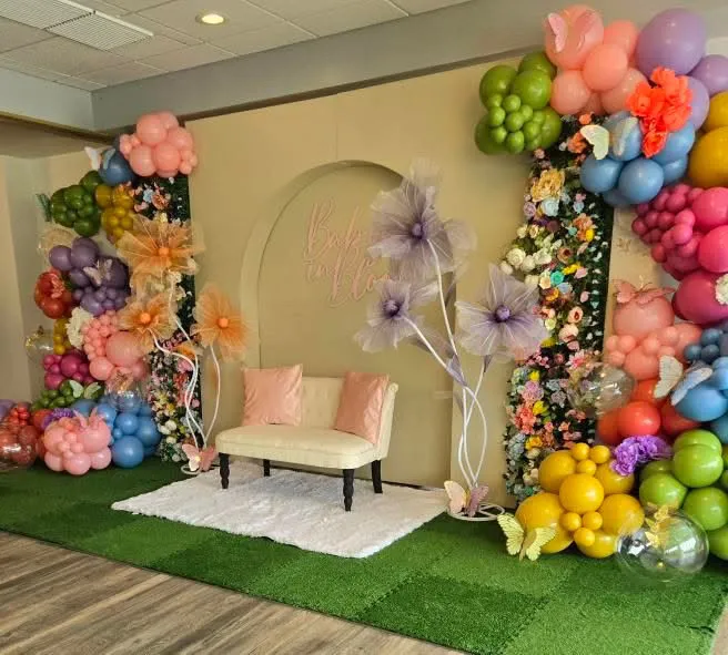 Baby shower Baby in Bloom backdrop with balloon garlands, floral arrangements, cream settee, and green turf flooring.