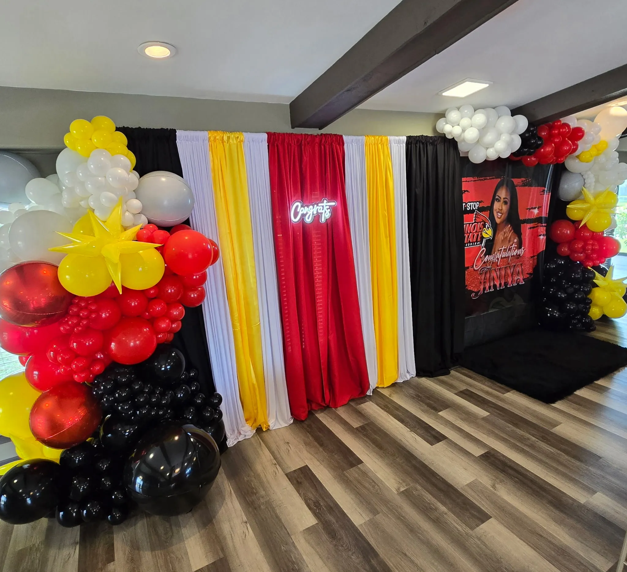 Graduation celebration backdrop with fabric drapes, balloon garlands, a Congrats neon sign, and a custom banner.