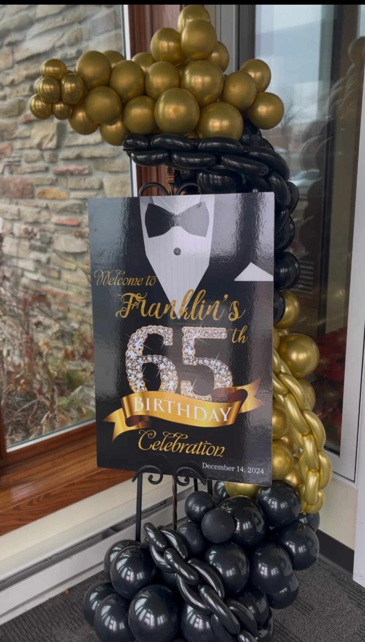 65th birthday welcome sign with a gold and black balloon garland, tuxedo graphic, and glitter accents.